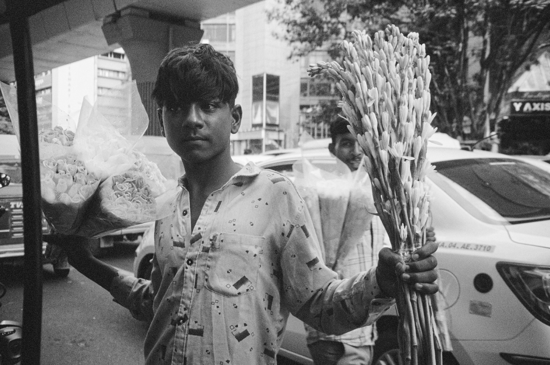 Black and white photograph of a flower seller with roses and tuberoses beside passing traffic